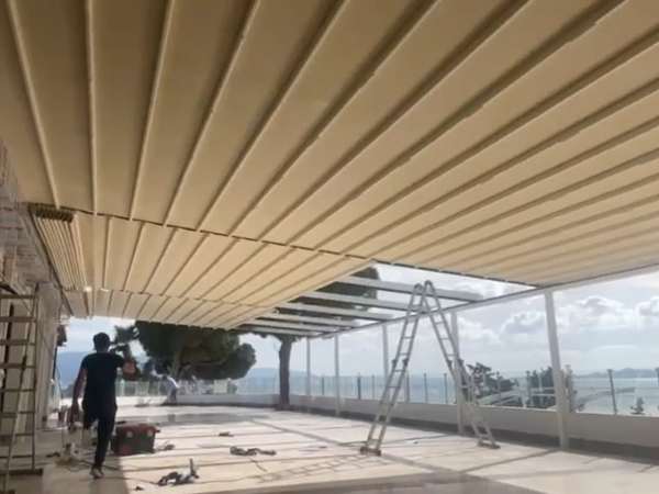 Antalya'da deniz manzaralı otel-kafe terasına kurulan krem renkli katlanır kumaş çatı sistemi; montaj sırasında alttan görünüm ve arka planda deniz