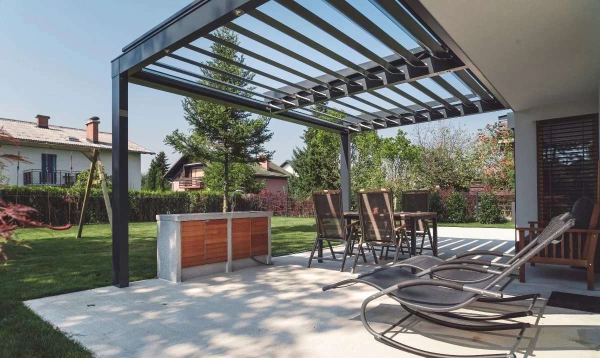 Villa bahçesindeki yemek alanına kurulan antrasit bioklimatik pergola; lameller kısmen açık, altında masa ve sandalyeler duruyor