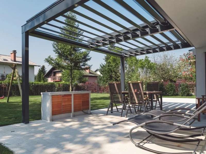 Villa bahçesindeki yemek alanına kurulan antrasit bioklimatik pergola; lameller kısmen açık, altında masa ve sandalyeler duruyor