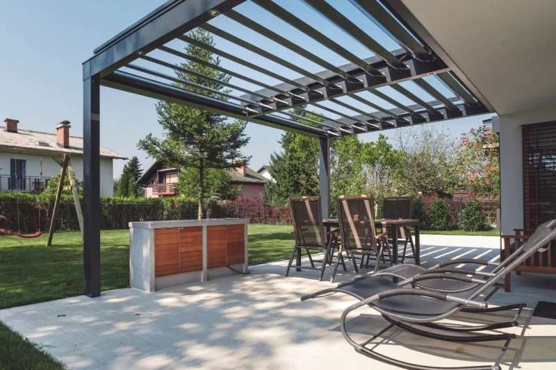 Villa bahçesindeki yemek alanına kurulan antrasit bioklimatik pergola; lameller kısmen açık, altında masa ve şezlonglar