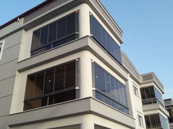Apartmanın üst iki katındaki köşe balkonları antrasit doğramalı füme camlı cam balkon sistemiyle kapatılmış; paneller kapalı konumda, sağda aynı sistemle kapatılmış komşu blok