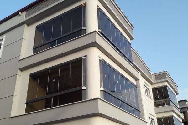 Apartmanın üst iki katındaki köşe balkonları antrasit doğramalı füme camlı cam balkon sistemiyle kapatılmış; paneller kapalı konumda, sağda aynı sistemle kapatılmış komşu blok