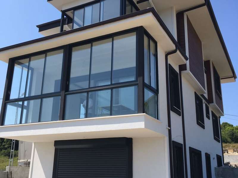 Müstakil evin köşe balkonu antrasit doğramalı cam balkon sistemiyle kapatılmış; camlar kapalı, alt katta aynı renkte sarmal kepenkli garaj kapısı ve henüz tamamlanmamış çevre düzenlemesi