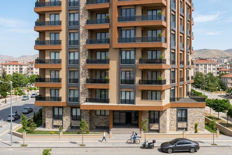 Çok katlı bir apartmanın köşe cephesi ahşap desenli kompozit panel ve taş görünümlü kaplamayla kaplanmış; antrasit alüminyum doğramalar, cam balkon korkulukları ve zemin katta taş kaplamalı giriş bölümü caddeden görünüyor