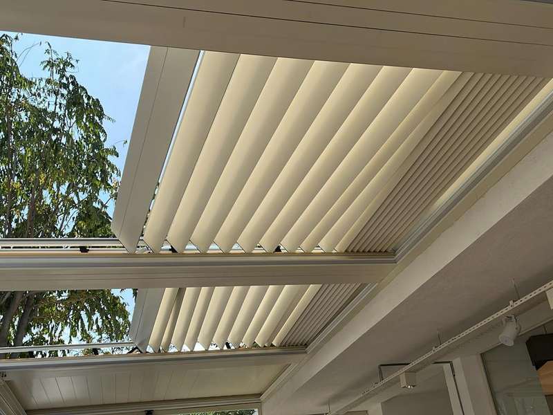 Yarı açık konumdaki krem rolling roof çatı; toplanan paneller arasından gökyüzü ve ağaç dalları görünüyor