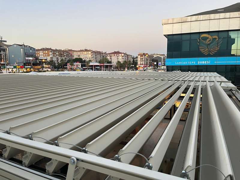 Sancaktepe şubesi binasının üstündeki beyaz rolling roof sisteminin dıştan üst görünümü; kapalı konumdaki paneller ve arkada şehir silueti