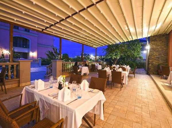 Akşam servisine hazırlanmış restoran terası: krem katlanır çatı, LED aydınlatma, beyaz örtülü masalar ve mavi ışıklı bina cephesi