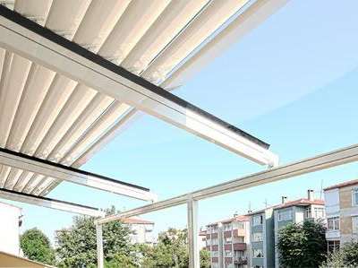 Yarı açık krem katlanır çatının alttan görünümü; açılan bölümden mavi gökyüzü ve arkada konut yerleşimi görünüyor