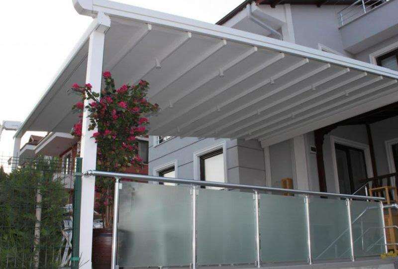 Beyaz katlanır çatının konut cephesindeki uygulaması; cam korkuluklu balkon, sardunya saksıları ve beyaz doğramalı pencereler