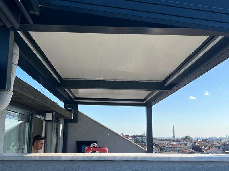 Çatı terasına kurulan antrasit taşıyıcılı beyaz katlanır çatının içeriden görünümü; arkada İstanbul silueti ve minare