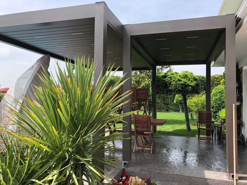 Yağmur sonrası bahçede antrasit bioklimatik pergola; ıslak zemin, altında ahşap masa ve sandalyeler