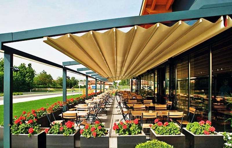 Uzun restoran terası boyunca açılmış krem katlanır çatı; sıralı ahşap masalar, çiçek saksıları ve cam cephe