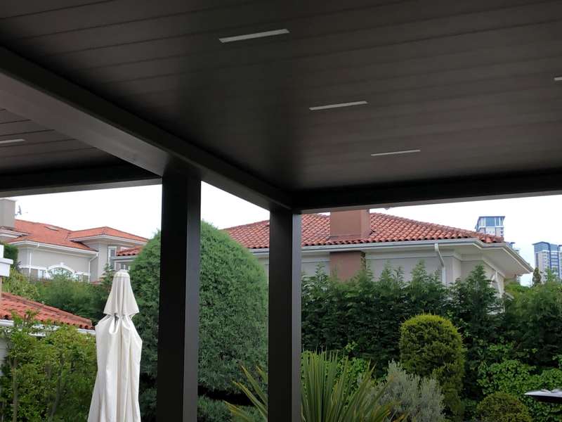 Site içindeki villa terasına kurulan antrasit bioklimatik pergola; kapalı lameller, kapalı şemsiye ve kiremit çatılı komşu evler