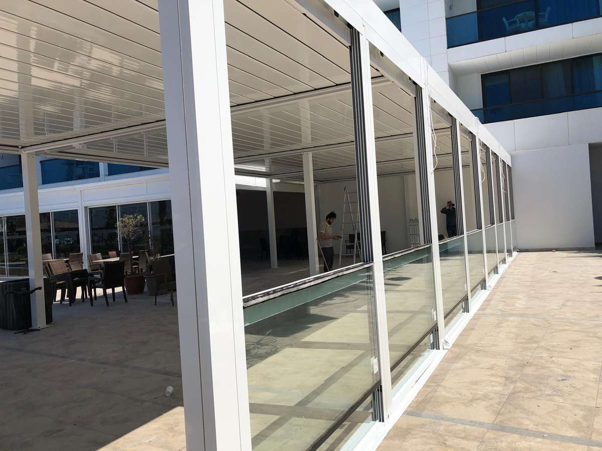 Otel terasına cephe boyunca kurulan beyaz doğramalı giyotin cam sistemi; alt paneller yarıya kadar kaldırılmış, üstte beyaz lamelli pergola çatısı ve solda teras oturma alanı