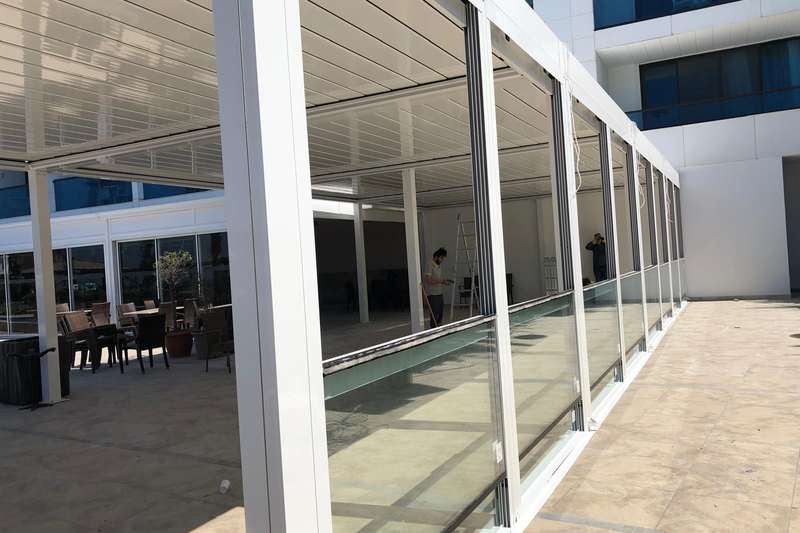 Otel terasına cephe boyunca kurulan beyaz doğramalı giyotin cam sistemi; alt paneller yarıya kadar kaldırılmış, üstte beyaz lamelli pergola çatısı ve solda teras oturma alanı