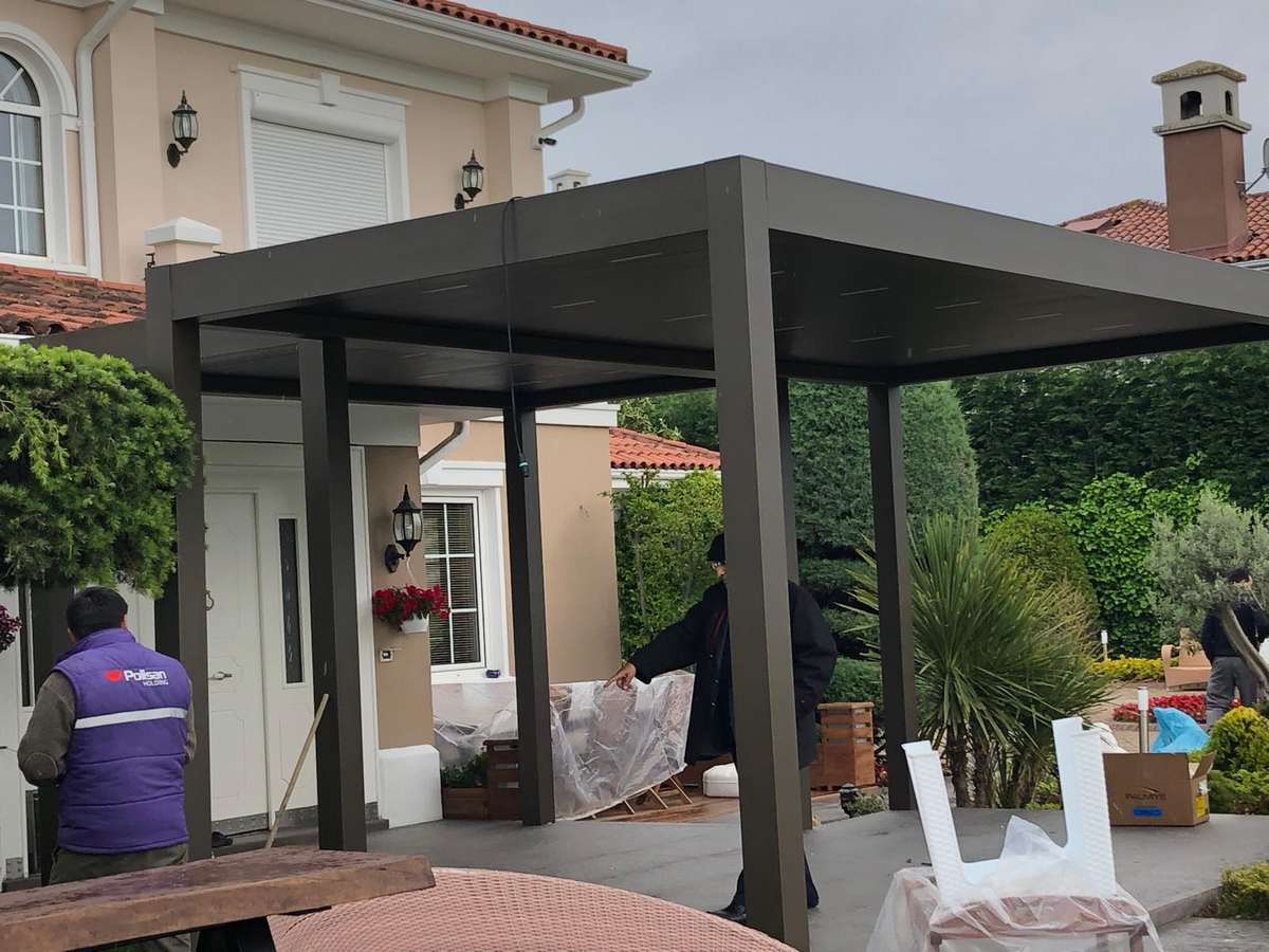Antalya'da villa girişinin önüne kurulan antrasit renkli rolling roof pergola; montaj ekibi çalışırken giriş kapısı ve bahçe görünüyor