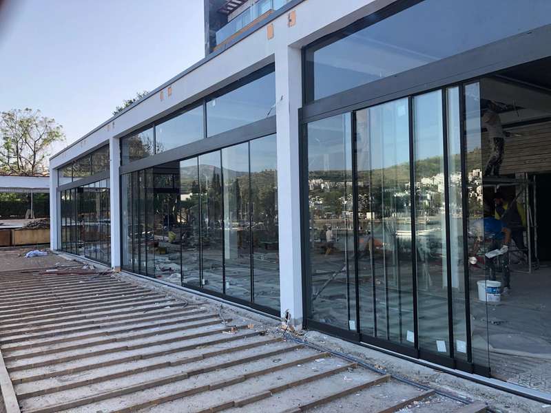 İşletme dış alanına kurulan beyaz pergola ve geniş modüllü sürgülü cam sistemi; montaj tamamlanma aşamasında, arka planda yerleşim görünüyor
