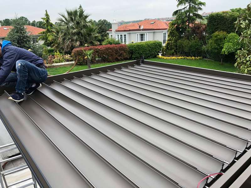 Bioklimatik pergola çatısının montaj sırasında üstten görünümü; ekip üyesi antrasit lamelleri yerleştiriyor, arkada site ve yeşil alan