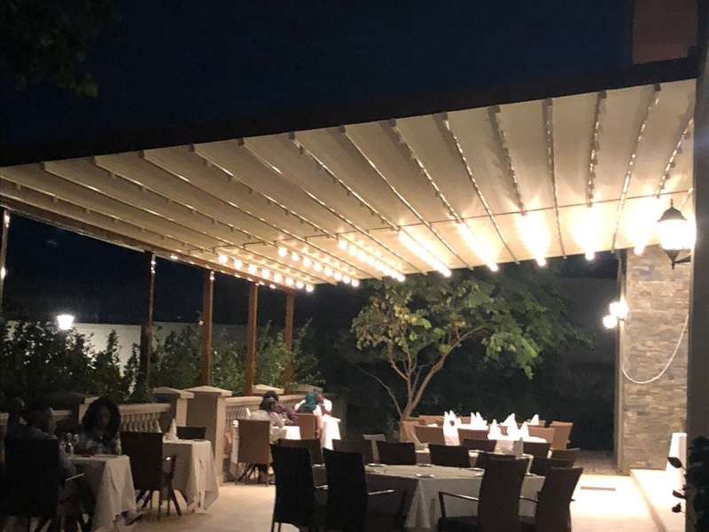 Akşam saatinde restoran terası: LED şeritli beyaz pergola çatısı, taş duvar ve oturmuş misafirler