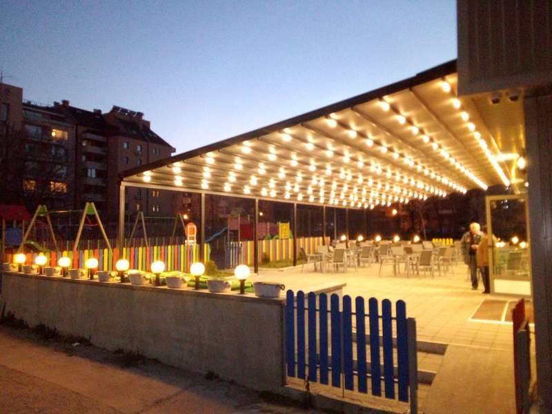 Sokak üzerindeki işletmenin önüne kurulan uzun pergola; LED aydınlatma, sarı-mavi bahçe çiti ve masa düzeni