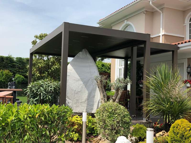 Villa bahçesindeki antrasit bioklimatik pergolanın montaj sonrası hâli; örtülü mobilya, zeytin ağacı ve krem cepheli ev