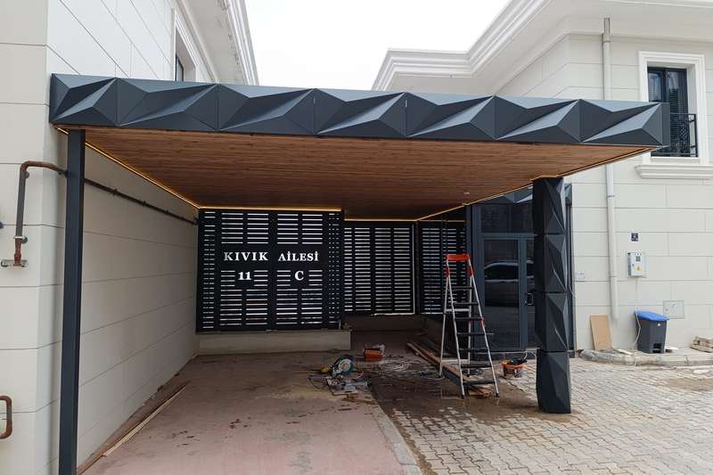 Villa girişine kurulan araç gölgeliği; alınlıkta üç boyutlu kırılımlı antrasit kompozit panel, tavanda ahşap desenli kaplama ve gömme LED şerit, arkada lazer kesim desenli bahçe kapısı