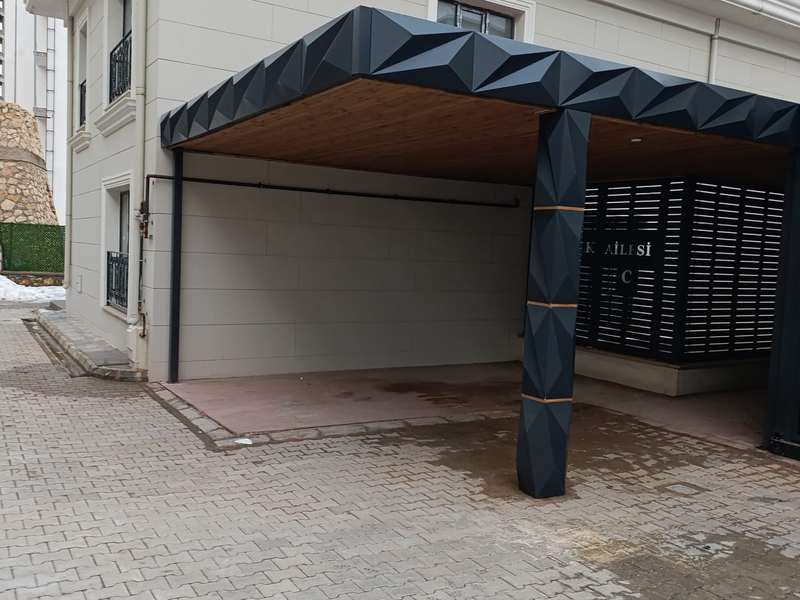 Tamamlanmış araç gölgeliğinin yandan görünümü; üç boyutlu kırılımlı panellerle giydirilmiş taşıyıcı kolon ve alınlık, altında kilit taşı döşeli park alanı