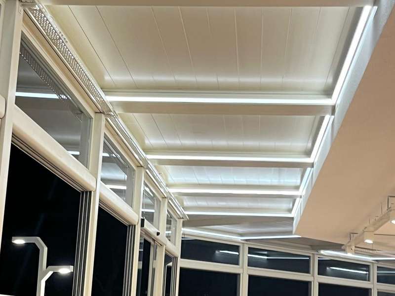 Kafe dış alanına kurulan beyaz lamelli pergola ve giyotin cam sistemi; içeriden çatı lamelleri ve kapatılmış cam cephe görünüyor