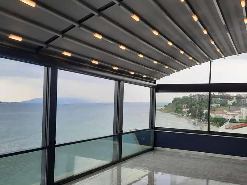 Deniz manzaralı terasa kurulan LED aydınlatmalı kavisli pergola ve giyotin cam sistemi; cam cephe boyunca kıyı ve deniz görünüyor