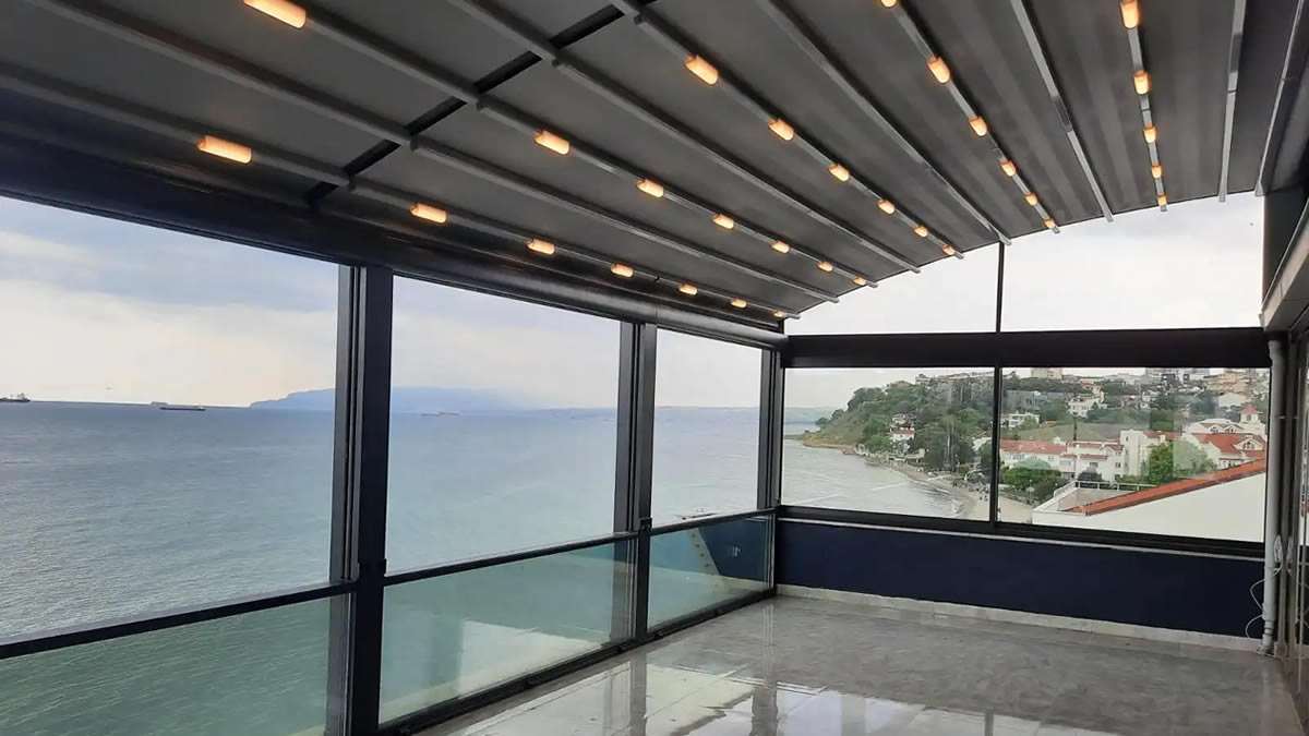 Deniz manzaralı terasa kurulan LED aydınlatmalı kavisli pergola ve giyotin cam sistemi; cam cephe boyunca kıyı ve deniz görünüyor