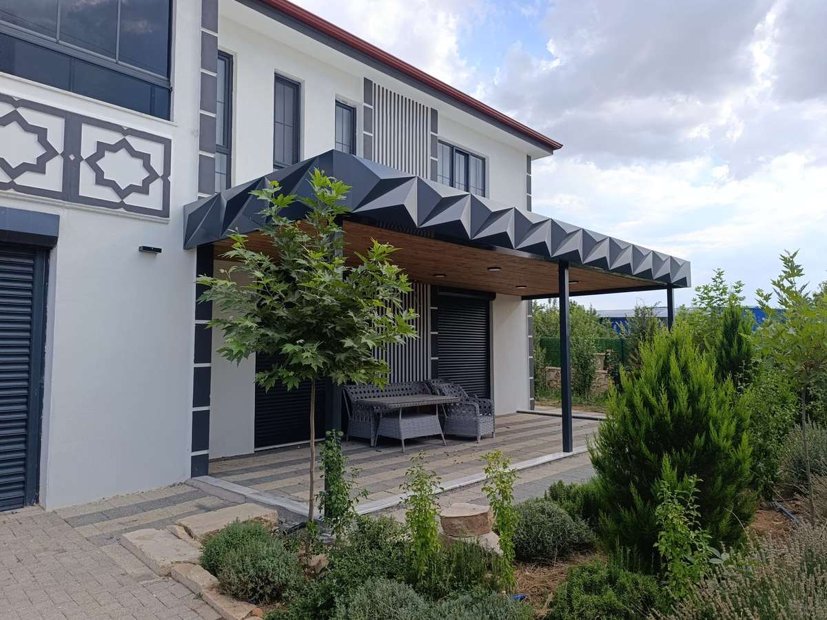 Villanın bahçe cephesine yaslanan antrasit veranda; alınlıkta üç boyutlu kırılımlı alüminyum kompozit panel, tavanda ahşap desenli kaplama ve gömme spotlar, altında rattan oturma grubu
