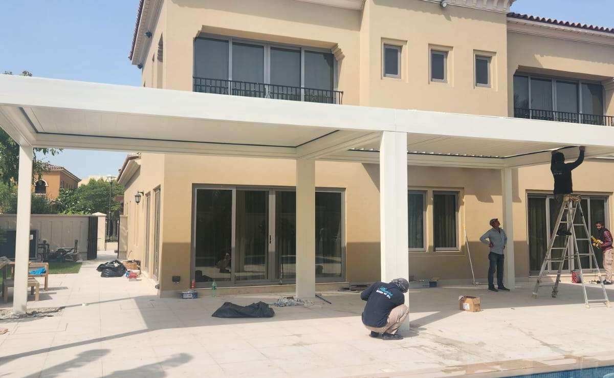Villa cephesine kurulan beyaz bioklimatik pergolanın montaj aşaması; taşıyıcı kolonlar dikilmiş, ekip lamel montajı yapıyor